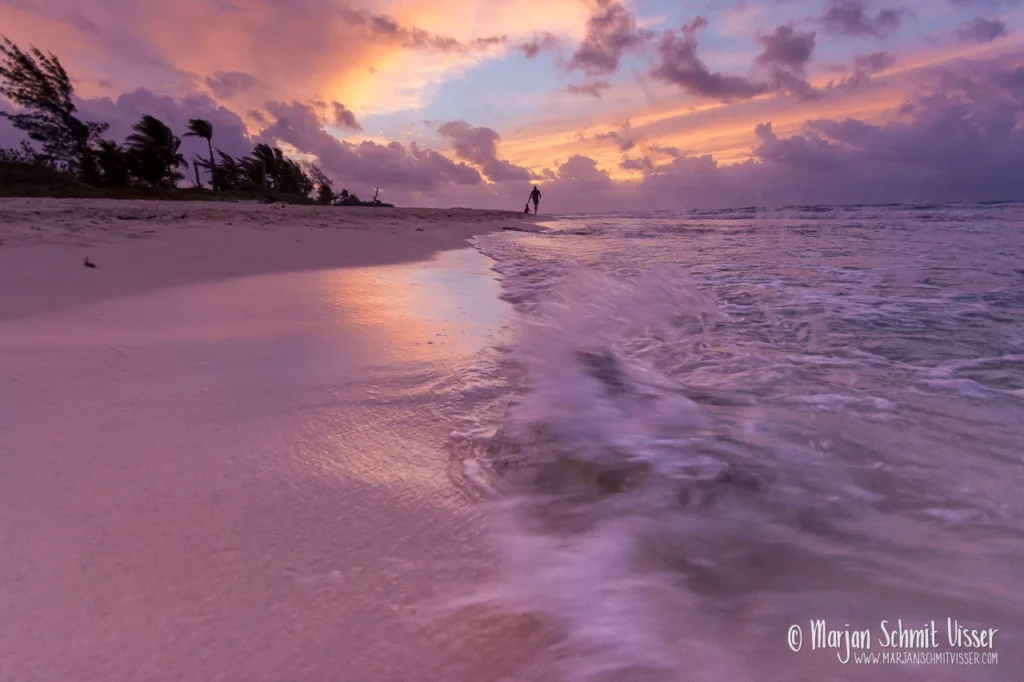 Zonsopkomst boven de zee bij Playa del Carmen, Mexico, met een wandelaar op het strand en golven die zacht over het zand spoelen.