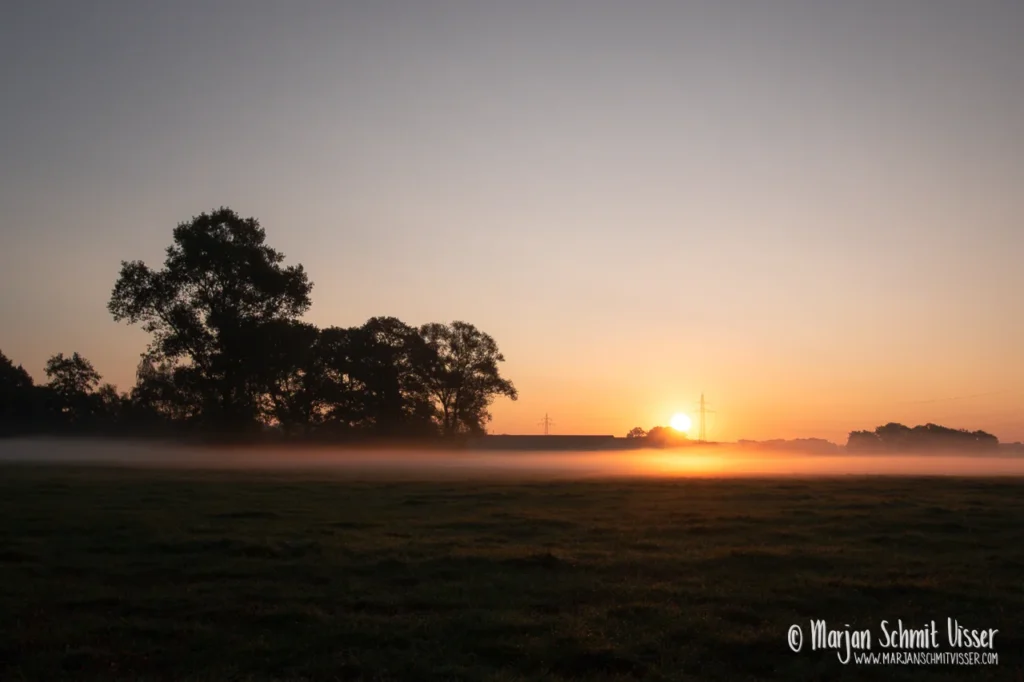 Zonsopkomst boven een nevelig veld bij Mariënheem in Nederland, met bomen en een gouden gloed aan de horizon.