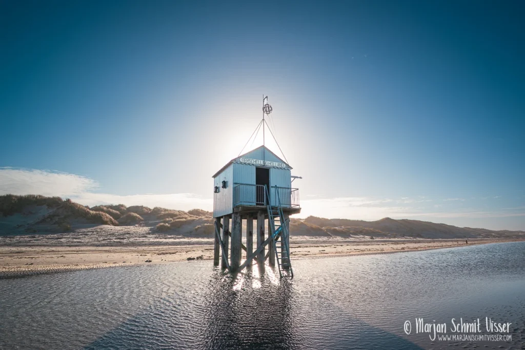 Drenkelingenhuisje op het strand van Terschelling, Nederland, in tegenlicht met zon achter het huisje en helderblauwe lucht.