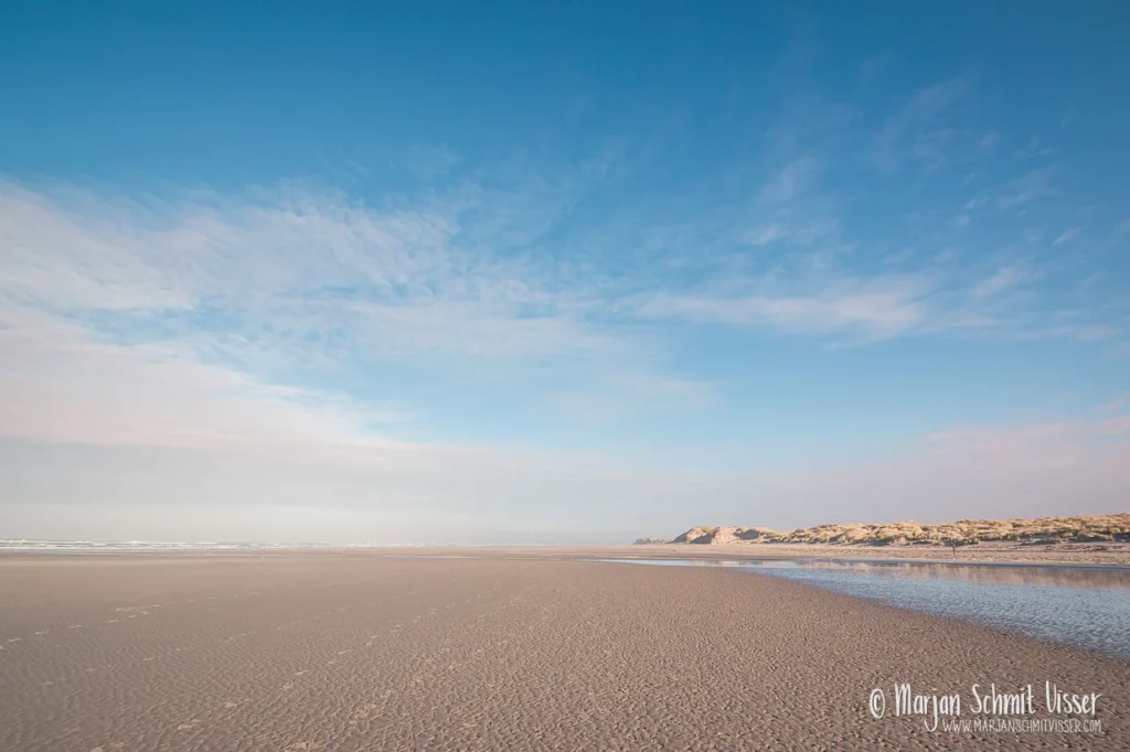 Uitzicht over het uitgestrekte strand van Terschelling, Nederland, met blauwe lucht, duinen in de verte en weerspiegeling van de zee.