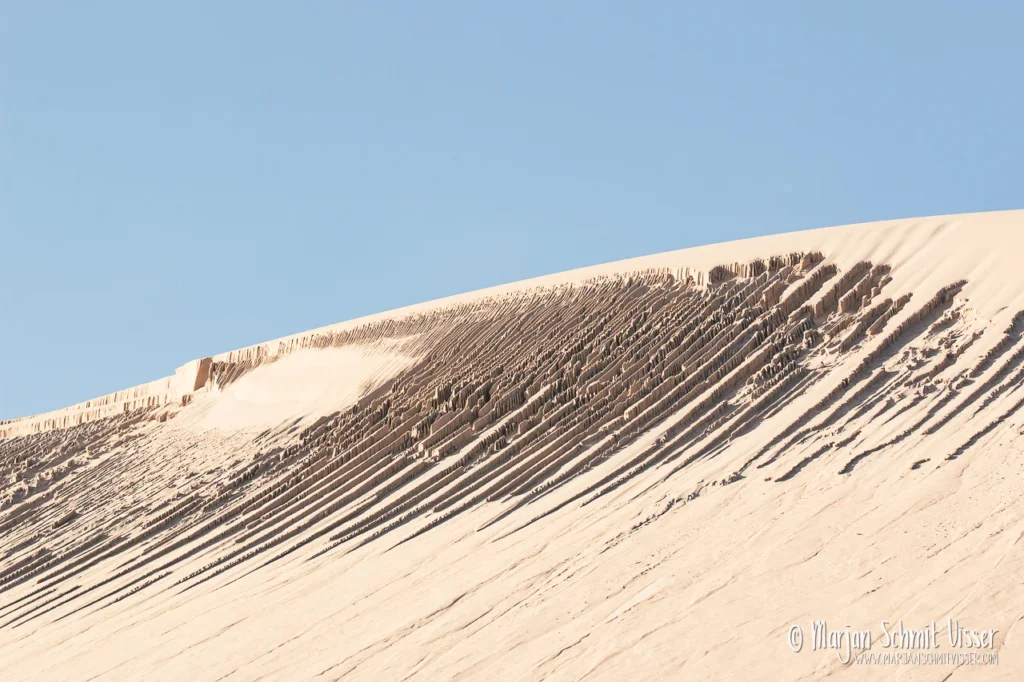 Aan de Nederlandse kust 2 Aan de Nederlandse kust Detail van een zandduin op Terschelling, Nederland, met windpatronen en gelaagde lijnen onder een heldere blauwe lucht.